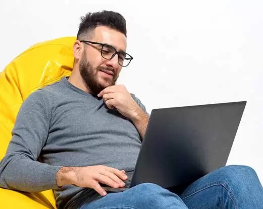 A man with a laptop on his lap, working comfortably on a couch in a home office setting.